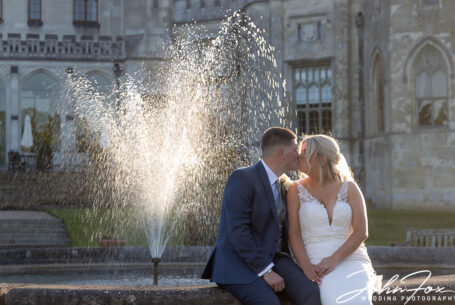 Bride and groom kissing by a back lit fountain in the sun at Ashridge House. Image by John Fox Wedding Photography.