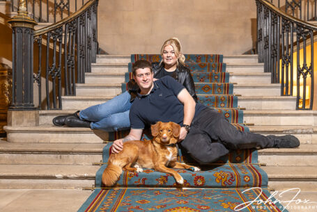 Bride and groom and their dog lounging on the main stairs of Ashridge House. Image by John Fox Wedding Photography.