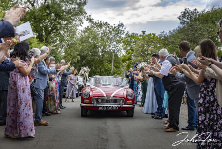 Wedding photographer in Oxfordshire. Bride and groom in wedding car leaving the church with guests waving.