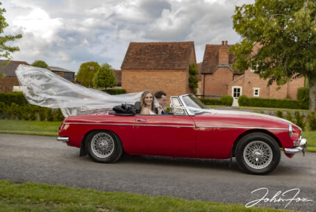 Bride with veil blowing in the wind as they drive away in an MG