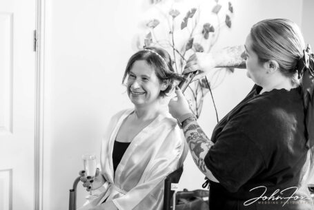 Rebecca the bride smiling and enjoying a glass of bubbly while having her hair prepared for the ceremony at Hadley Park House