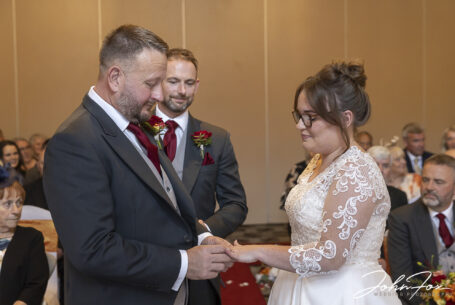 Wedding ceremony ring exchange; Matthew putting the ring on Katie's finger with the best man looking on.