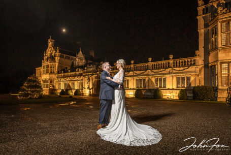 Joanne and Kevin, the bride and groom, outside Rushton Hall after dark with the Hall lit up behind them.