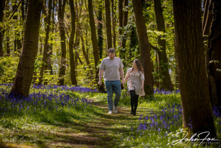 An image of the happy couple, Rikki and Jack, walking amongst the trees and bluebells. Pre-wedding shoot in Warwickshire woodland. Wedding Photographer in the West Midlands, John Fox.