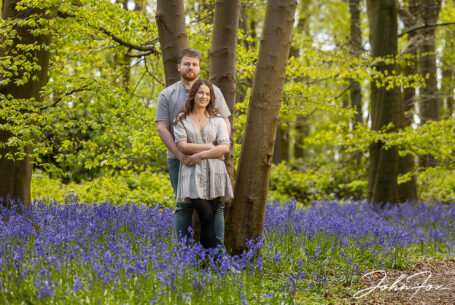 An image of the happy couple, Rikki and Jack, holding each other while leaning against a tree with bluebells all around them. Pre-wedding shoot in Warwickshire woodland. Wedding Photographer in the West Midlands, John Fox.