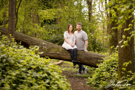 An image of the bride-to-be, Rikki, sitting on a fallen tree with Jack standing by her. Pre-wedding shoot in Warwickshire woodland. Wedding Photographer in the West Midlands, John Fox.