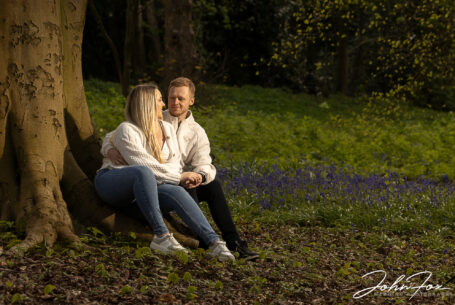 Pre-wedding shoot at Mapperley Reservoir woodland area with happy couple sat by a tree and looking lovingly at each other.
