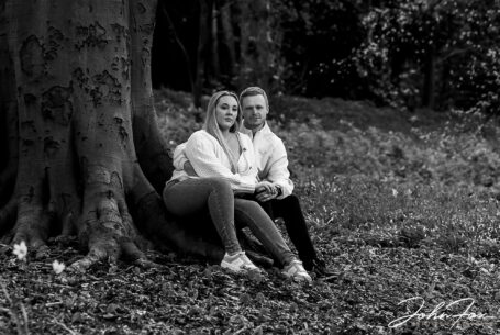 Pre-wedding black and white image at Mapperley Reservoir's woodland area with the happy couple sat by a tree holding hands